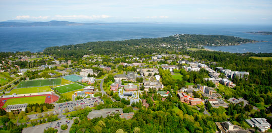 Aerial view of UVic's campus, photo taken on a sunny day. The ocean is visible in the distance. 